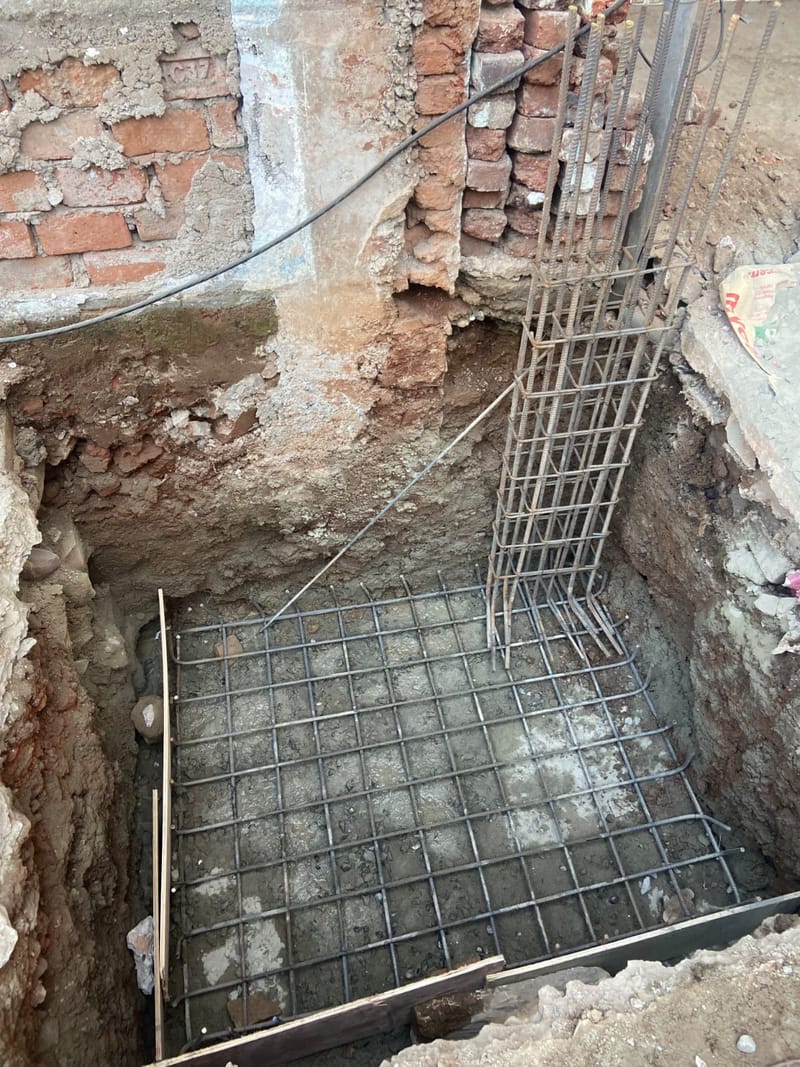 Excavated footing pit with a steel reinforcement mesh on the levelling bed and a vertical column cage, beside an old brick wall.